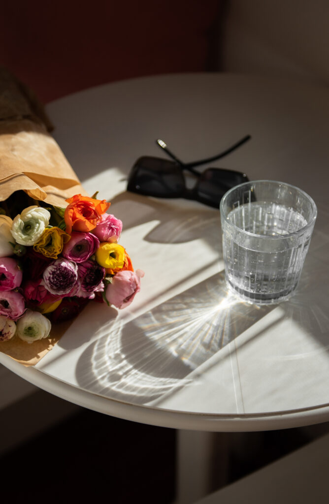 Bouquet of colorful flowers beside a glass of sparkling water and sunglasses in warm sunlight.
