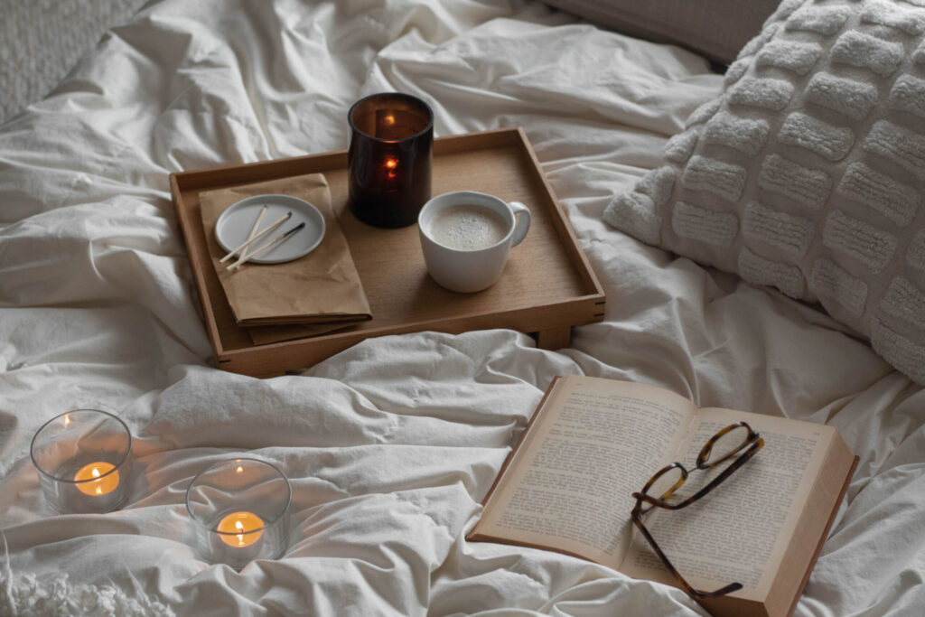 Wooden bed tray with coffee, lit candle, matches, and an open book on soft white bedding.
