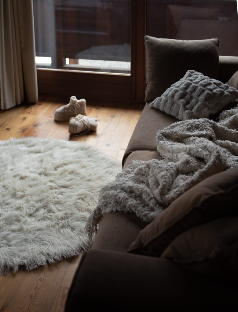 Soft sofa with textured pillows and blanket beside a fluffy rug and slippers in a warm cabin setting.