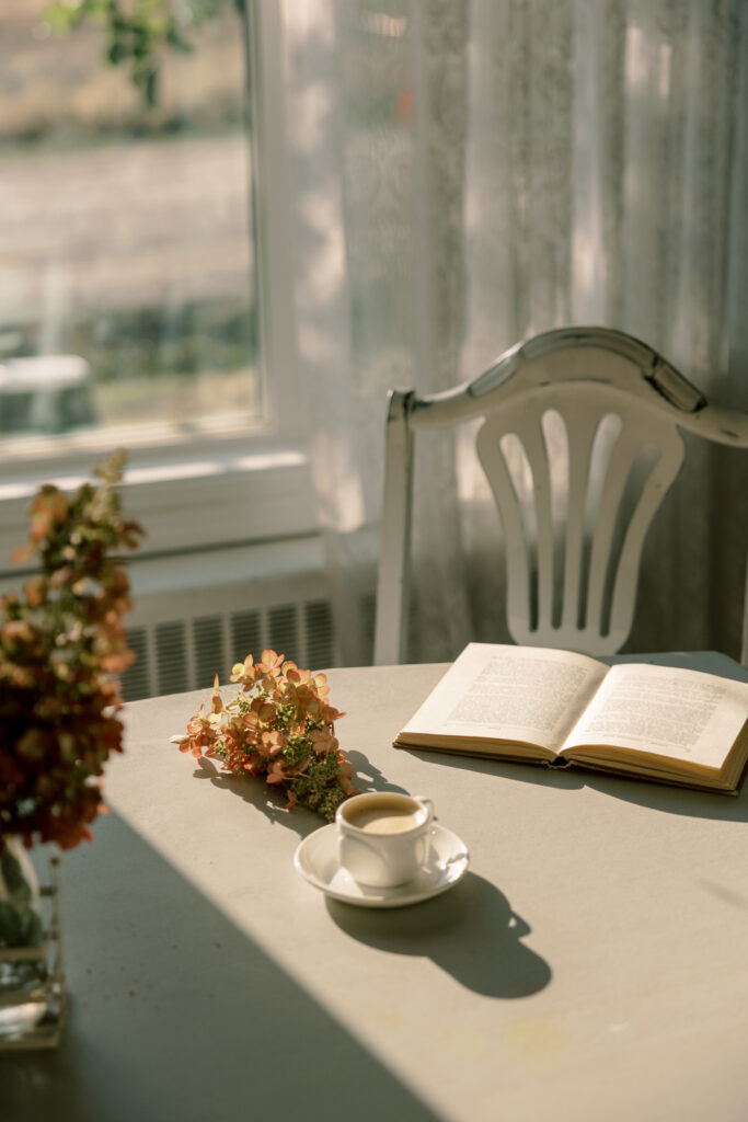 Open book and coffee cup on a table near a window with soft afternoon sunlight.