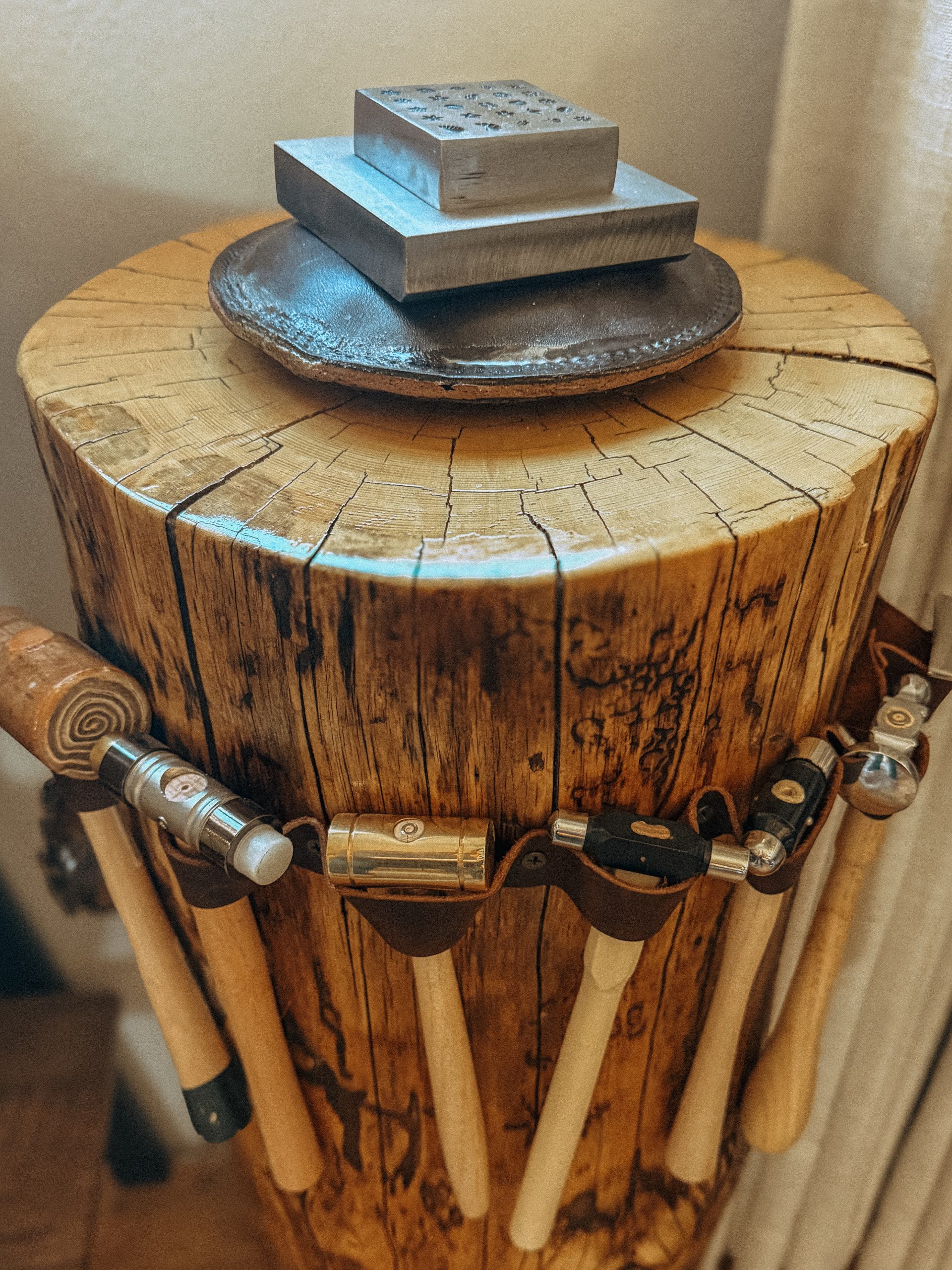 A collection of silversmithing hammers and a steel block resting on a wood stump in the Foreverie jewelry studio.