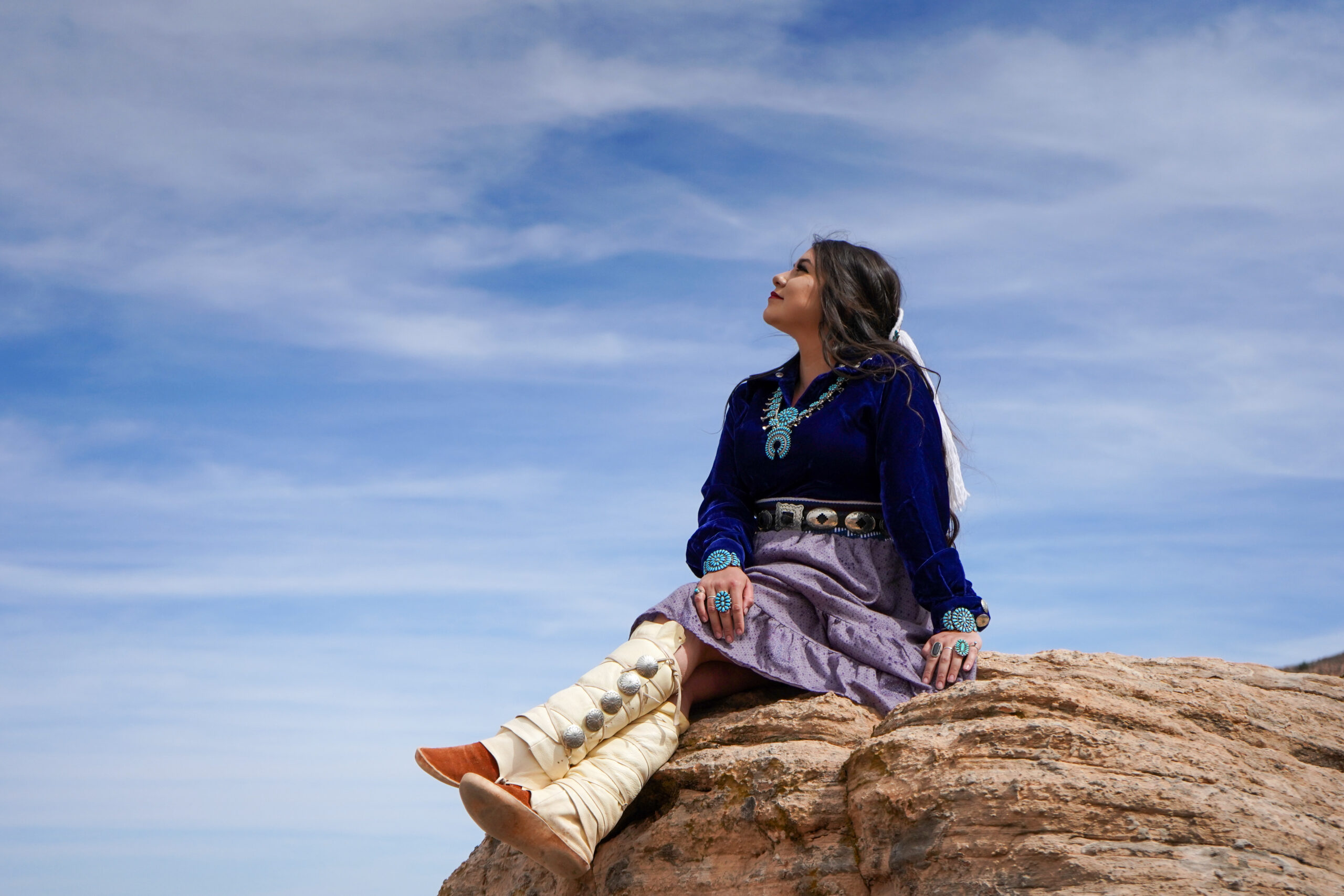 Kayla Perry wearing traditional Navajo clothing and turquoise jewelry, seated on red rock under a bright desert sky.
