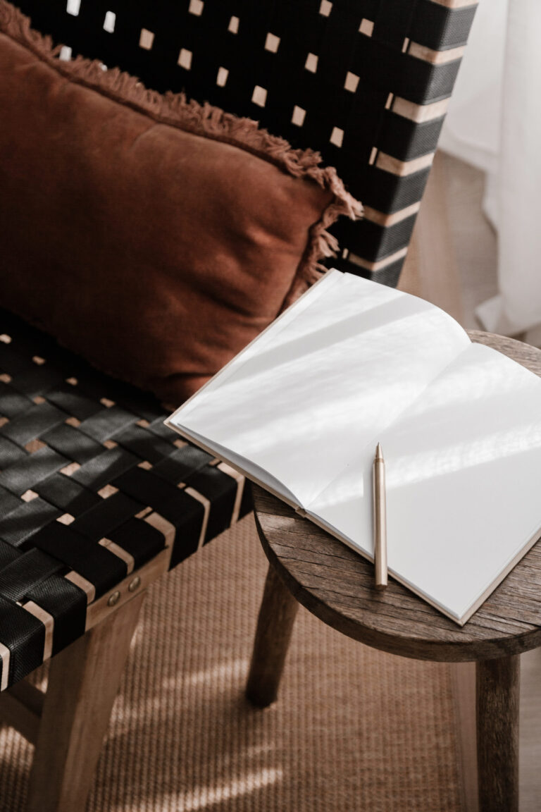 Open blank journal with pen on a wooden stool beside a woven chair in soft morning light
