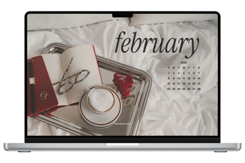Laptop mockup displaying February 2026 desktop wallpaper with coffee, notebook, and silver tray on white bedding.