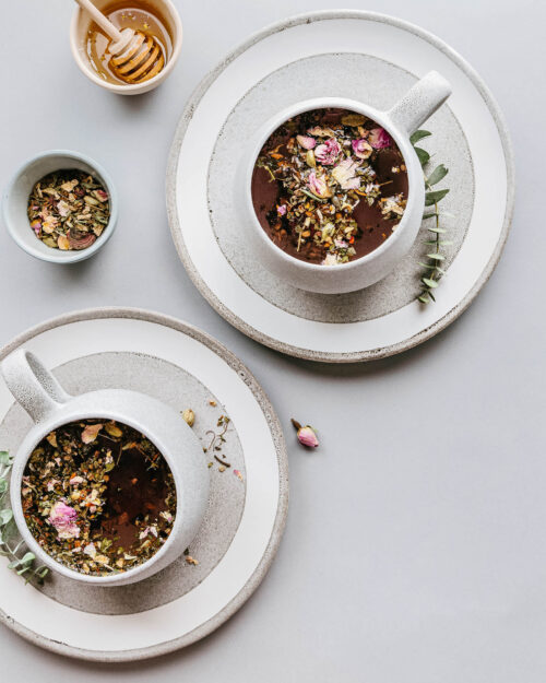 Two cups of herbal tea topped with dried flowers, styled with honey and loose herbs on neutral plates.