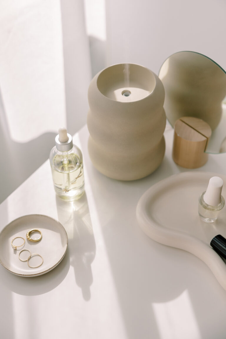 Neutral vanity with skincare serum, gold rings, nail polish, and sculptural diffuser in natural light.