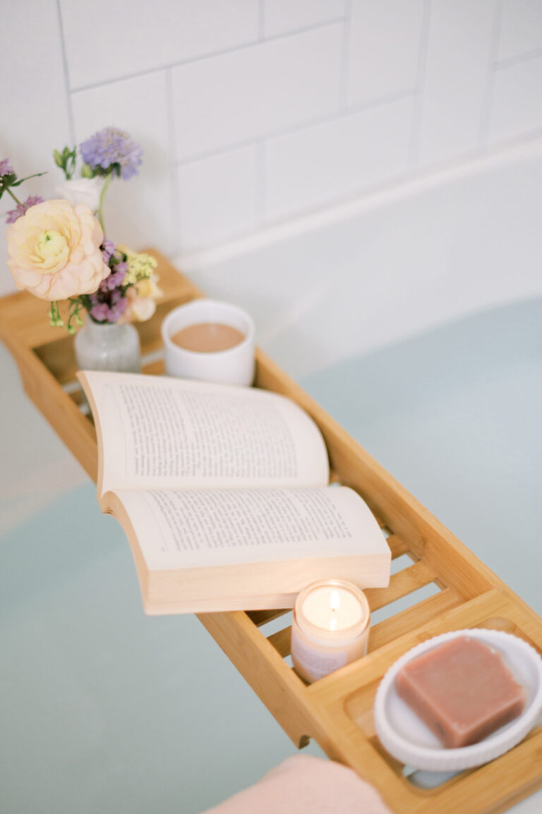 Wooden bath tray with an open book, lit candle, flowers, tea, and soap over a bathtub.