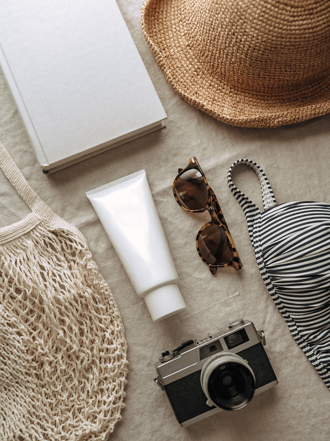 flat lay of travel essentials including woven bag, sunglasses, film camera, sunscreen, and striped swimwear