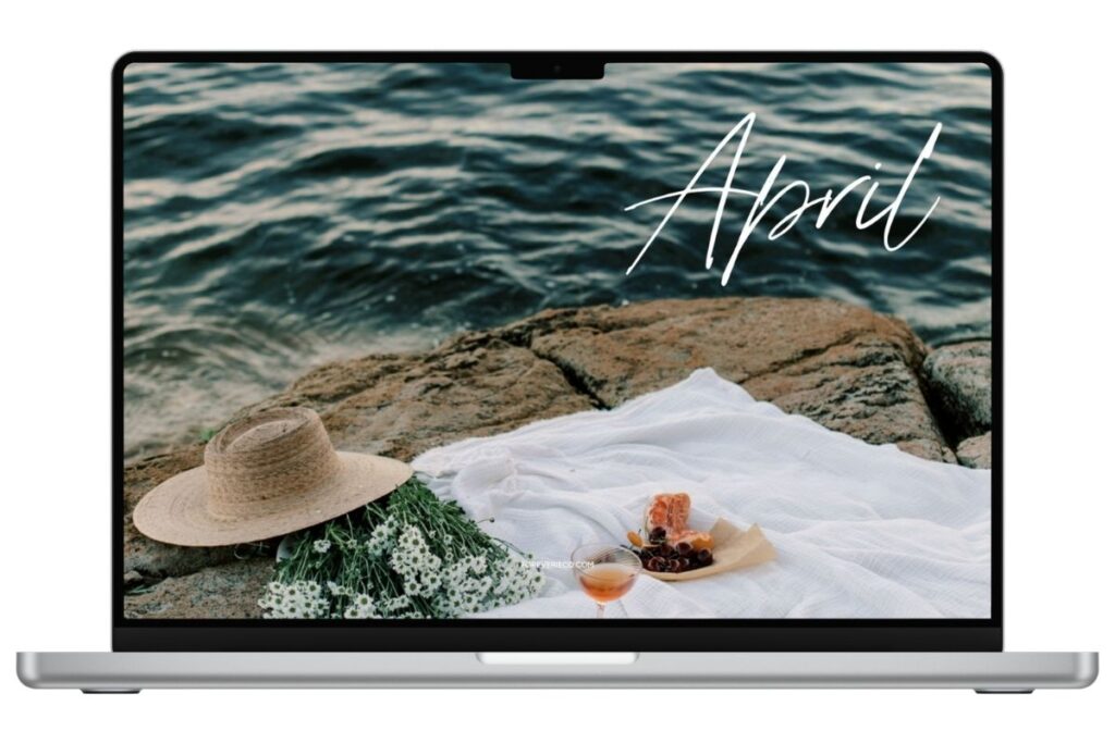 Laptop mockup displaying April 2026 desktop wallpaper with coastal picnic scene by the ocean