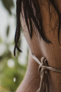 Close-up of a person with wet hair and water droplets on their skin wearing a neutral swimsuit outdoors.