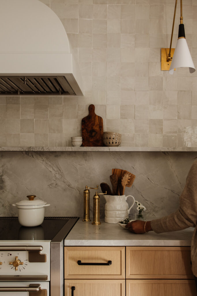 Warm neutral kitchen with creamy tile backsplash, marble countertops, brass pepper mills, wood utensils, white ceramic cookware, and light oak cabinets.