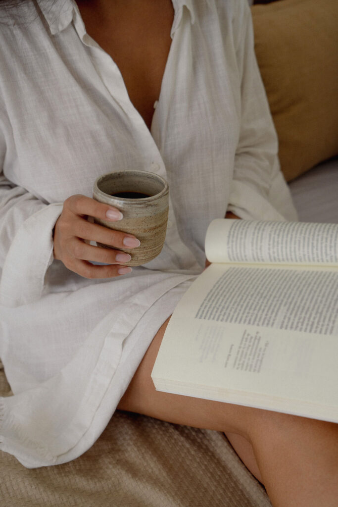 A person in a white linen shirt holds a rustic ceramic mug while reading an open book, creating a serene and cozy atmosphere.